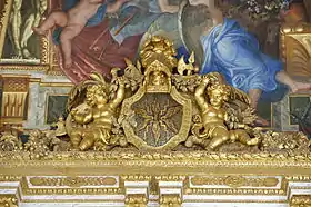 Photographie de la jonction entre les murs et le plafond. Un trumeau doré avec des angelots encadrant un écu où sont représentés les insignes de Jupiter : la foudre et les éclairs