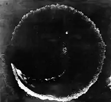 Photograph looking directly down at the ocean's surface from a high altitude. There is a rough circle on the surface that is about as wide as the photograph. The top deck of a ship is seen at the lower left of the circle; the length of the ship is much smaller than the circumference of the circle.