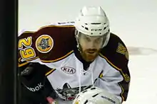A Caucasian ice hockey player bent over at the waist. He wears a white helmet and has a large beard.