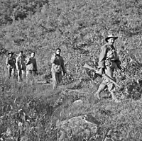 Three unarmed soldiers being led down a hill by two soldiers armed with rifles, one in the front the other in the rear.