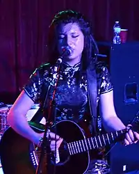 Chloe Chaidez performing on stage; singing into a microphone and playing a guitar