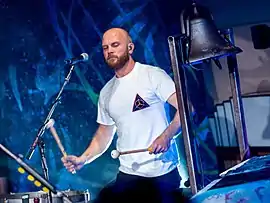 A bald, bearded man wearing a white shirt plays the timpani while a church bell is beside him