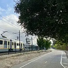 E Line train passes Expo Line Bikeway in Culver City; Hayden Tract architectural landmark visible in distance, native oak tree overhangs the path