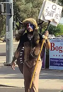 Chansley in his shaman dress on a sidewalk, holding a "Q sent me" sign