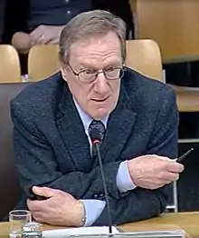 seated middle-aged White male wearing suit, speaking into microphone with crossed arms