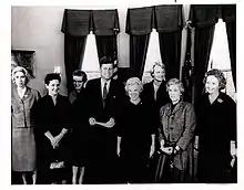 Seven women and President Kennedy standing in the Oval Office in 1962.