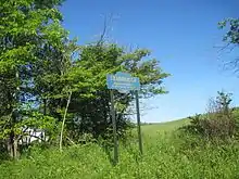 Image of a blue sign with gold writing supported by two green posts against a background of greenery. The sign reads, "STARRUCCA/FROM THE INDIAN/'JUNCTION OF WATERS'/FOUNDED/1818."