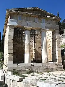 A partially ruined marble building with a porch with 2 columns supporting a pediment and an open doorway beyond