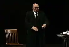 James Earl Jones standing on a stage with a black background with a wooden chair to his right and a table to his left while performing Driving Miss Daisy at the Theatre Royal in Sydney in March 2013