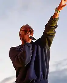 A black man with a blue sweater, orange glasses, and bleached dreadlocks singing into a microphone