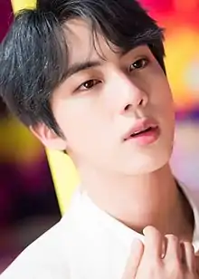 Kim in a white suit against a yellow purple and red bokeh background, he looks upwards and right