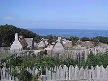 A modern day photograph of a village consisting of small, primitive wooden houses. Most of the houses have thatched roofs. In the distance is a large expanse of ocean and a clear blue sky. The village is surrounded by a wall consisting of tall, thick wooden planks.