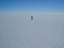 A panorama of flat, white ground, stretching to the horizon; a single person stands in the middle of the frame in the distance