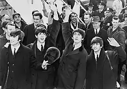 A black-and-white image of Paul McCartney, George Harrison, John Lennon and Ringo Starr waving to fans after arriving in America in 1964. A crowd is visible behind them on the left.