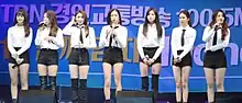 The members of CLC, in white shirts and black shorts, stand on a stage