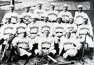 Baseball players are posing for a photograph, six men standing, seven men sitting on chairs, and three are sitting on the ground.