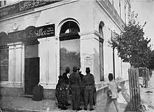 Photo of large white building with one sign saying "Moritz Schiller" and another in Arabic; in front is a cluster of people looking at a poster on the wall.