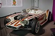 A white car with brown stripes, with open wheels and a clear bubble canopy over twin seats, and exposed, chromed engine with a blower. Beatnik Bandit II and a few of Roth's other cars are also on display in this museum.