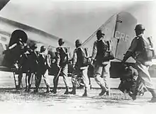 Men wearing parachutes and helmets file into a military aircraft on the ground