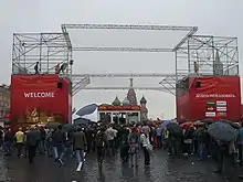 Two large red obelisks supporting a metal gantry. People with umbrellas are passing underneath the gantry.