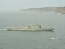 A large grey warship underway. Flags are being flown from the ship's mast, and white-uniformed people line the decks. Brown cliffs line the horizon for the right two-thirds of the image