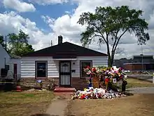 The single-story house has white walls, two windows, a central white door with a black door frame, and a black roof. In front of the house there is a walkway and multiple colored flowers and memorabilia.