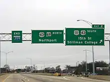 Three green signs are located above an elevated portion of roadway with no traffic visible on a cloudy day.