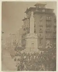 An antique photograph depicting a city square with a stone monument and a large number of soldiers at rest