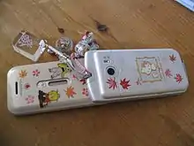 A Japanese flip phone typical of the 2000s with phone charms. These phones and charms were symbolic of youth culture in the early Heisei era, and remained prevalent in Japan long after the advent of the smartphone made flip phones obsolete elsewhere.