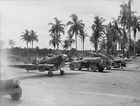 Black-and-white photo of two single-engined monoplane aircraft and a fuel tanker on a dirt clearing. Tropical palms are visible in the background.