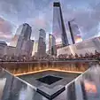 Illuminated water falls into the square 9/11 Memorial south pool at sunset, and glass-clad One World Trade Center and other skyscrapers rise in the background