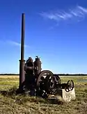 Photo of a very rusted industrial-looking pump with tall tube and wheel sits in a flat rice field.