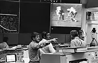 A control room; visible on a large screen are two astronauts walking on the Moon