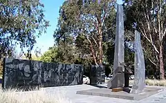 Monument consisting of two upright dark stones in front of a dark stone wall, with trees in the background