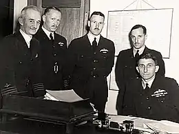 Five men, four of whom are standing and one seated, wearing dark-coloured military uniforms