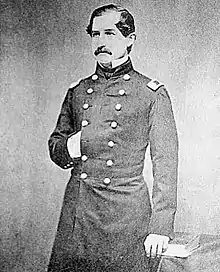 A three-quarters length monochrome photograph of a white man in a dark military uniform; he has dark hair and mustache, his right hand is tucked into the breast of his uniform, and he is looking to the camera's left.