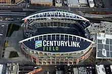 Aerial view of an empty stadium with prominent arches. The surrounding area has railroad tracks, a plaza, and a parking garage.