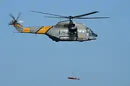 Spanish Air Force Aérospatiale SA330J Puma of 801 Squadron flying in an airshow. It is lifting a stretcher with a hoist. On the side of the helicopter is lettering reading "SAR", in yellow against the military grey colour scheme.