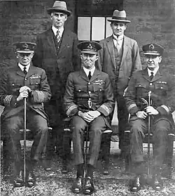 Formal portrait of three men in military uniforms with peaked caps sitting in front of two men in civilian clothes with broad-brimmed hats