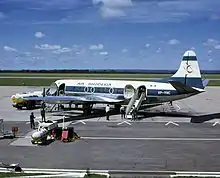 A white and blue aeroplane on a runway, surrounded by tankers of fuel and other equipment. The tail bears a stylised red Zimbabwe Bird, and the words "AIR RHODESIA" are painted above the windows of the cabin. Towards the rear are painted the letters "VP-YNC", with the green and white Rhodesian flag rendered above.