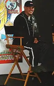 Akira Toriyama sitting on a chair smiling wearing black clothing with glasses and chin facial hair