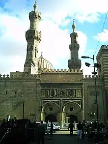 Two arched entrance-ways in the portico of a large two-story building face a street. Above the arches the building's wall is carved and ornamented. To the right, the building rises to a third story. Behind the wall two minarets framing the top of a dome are visible.