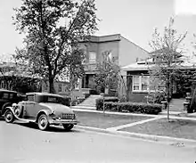 Two-story house with cars parked on the street in front of it.  It is partially obstructed by a neighboring house on the right.  A tall tree is on the lawn in front of the house.