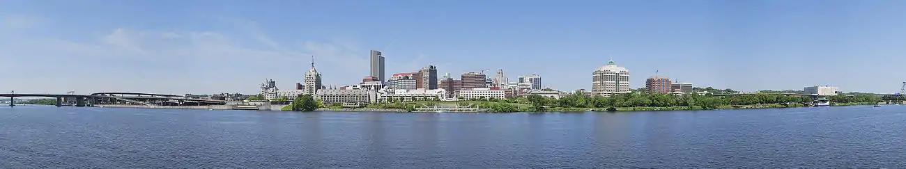 A panorama shows a river in the bottom half, crossed by a highway bridge on left; building towers are seen around the center, where a green zone on the bank of the river is seen, which extends to the right extreme of the image.