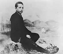 Studio photo of a boy seated in a relaxed posture and wearing a suit, posed in front of a backdrop of scenery.