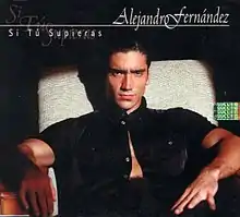 A man wearing an unbuttoned black shirt is sitting on a chair facing the camera