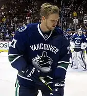 A Caucasian ice hockey player skating with gripping a hockey stick with both hands. He wears a green jersey with white and blue trim and a blue, visored helmet.