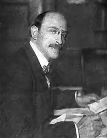 A 42-year-old man wearing a bowtie and suit leans over a desk as he writes