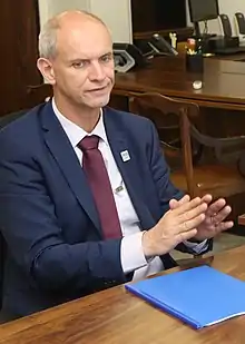 Middle-aged man with short hair speaking during a meeting. He is wearing a dark blue suit, with a white shirt and wine-colored neck tie.