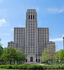 A tall white blocky stone building seen from across a square with trees and a walkway leading to the arched main entrance. Just below the top are three similar long narrow arched windows; the building has two wings on either side, about two-thirds its height, which themselves have wings about half their height from which American flags fly on poles on top.